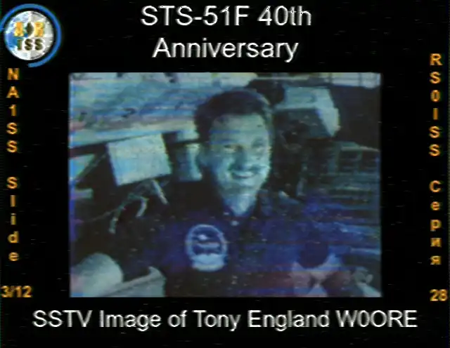 SSTV image from the ISS, Slide 3 of 12 40th Anniversary. The image is a bust photot of astronaut Tony England, who is smiling and looking towards the camera. He is wearing a blue polo shirt with a mission patch on the chest. The background shows the interior of the shuttle. The text at the bottom identifies the image as 'SSTV Image of Tony England W0ORE'. The image has a slight blueish tint