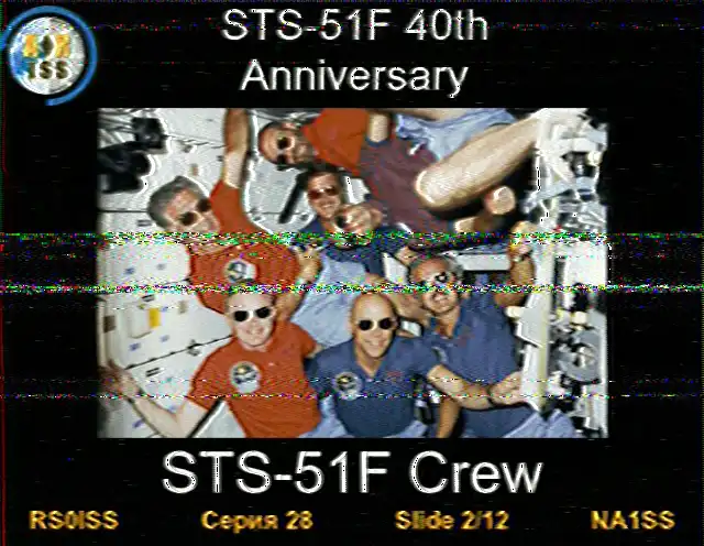 SSTV image from the ISS, Slide 2 of 12. The image features the seven-member crew of the Space Shuttle Challenger mission, floating together in the microgravity environment of the Spacelab module. All the astronauts are wearing sunglasses and smiling for a group photo. The text at the bottom reads 'STS-51F Crew'