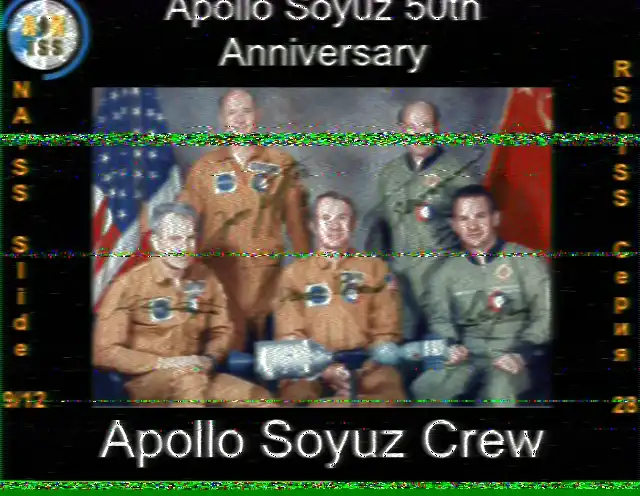 SSTV image from the ISS, Slide 9 of 12, celebrating the 50th anniversary of the Apollo-Soyuz mission. The image is a formal group portrait of the American astronauts and Soviet cosmonauts. The crew members are posed together in front of the American and Soviet flags, holding a model of the docked Apollo and Soyuz spacecraft. The text at the bottom reads 'Apollo Soyuz Crew'